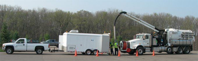 A white pickup truck towing a trailer, next to a large truck with a long arm, all parked on pavement.