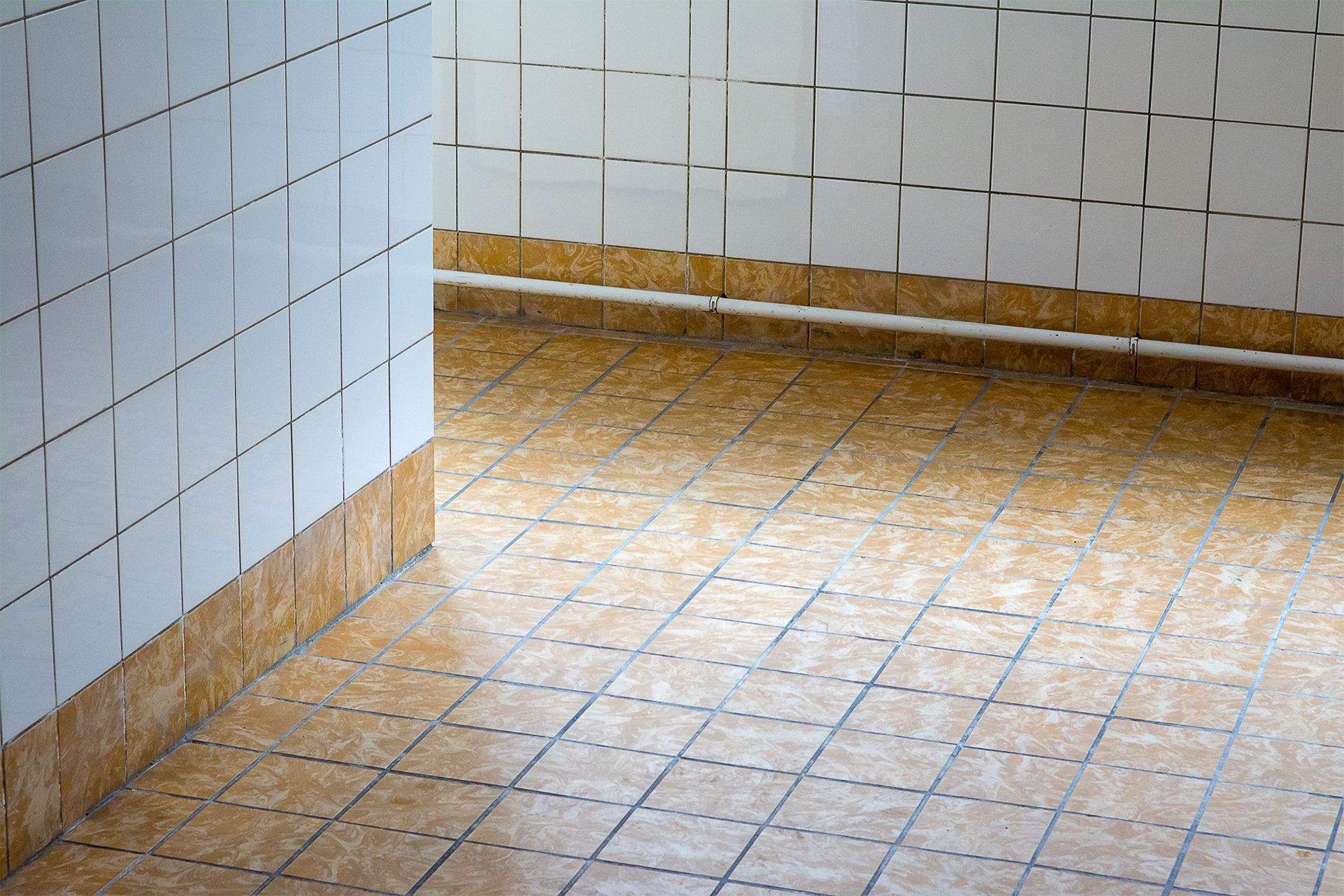 Empty tiled room corner with white walls and tan patterned floor tiles.