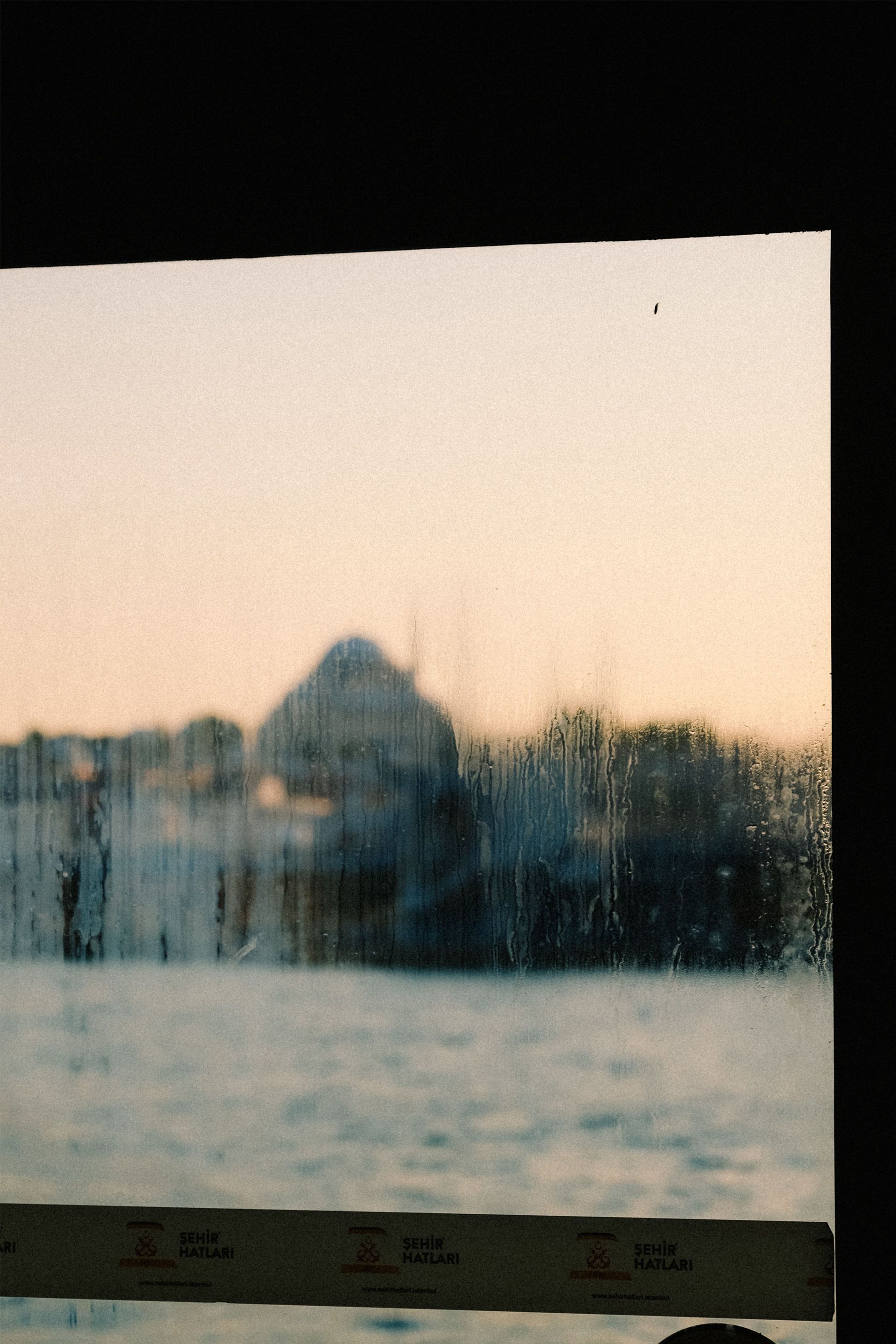 Snowy landscape at dusk with a distant building and bare trees beyond a window with water droplets.