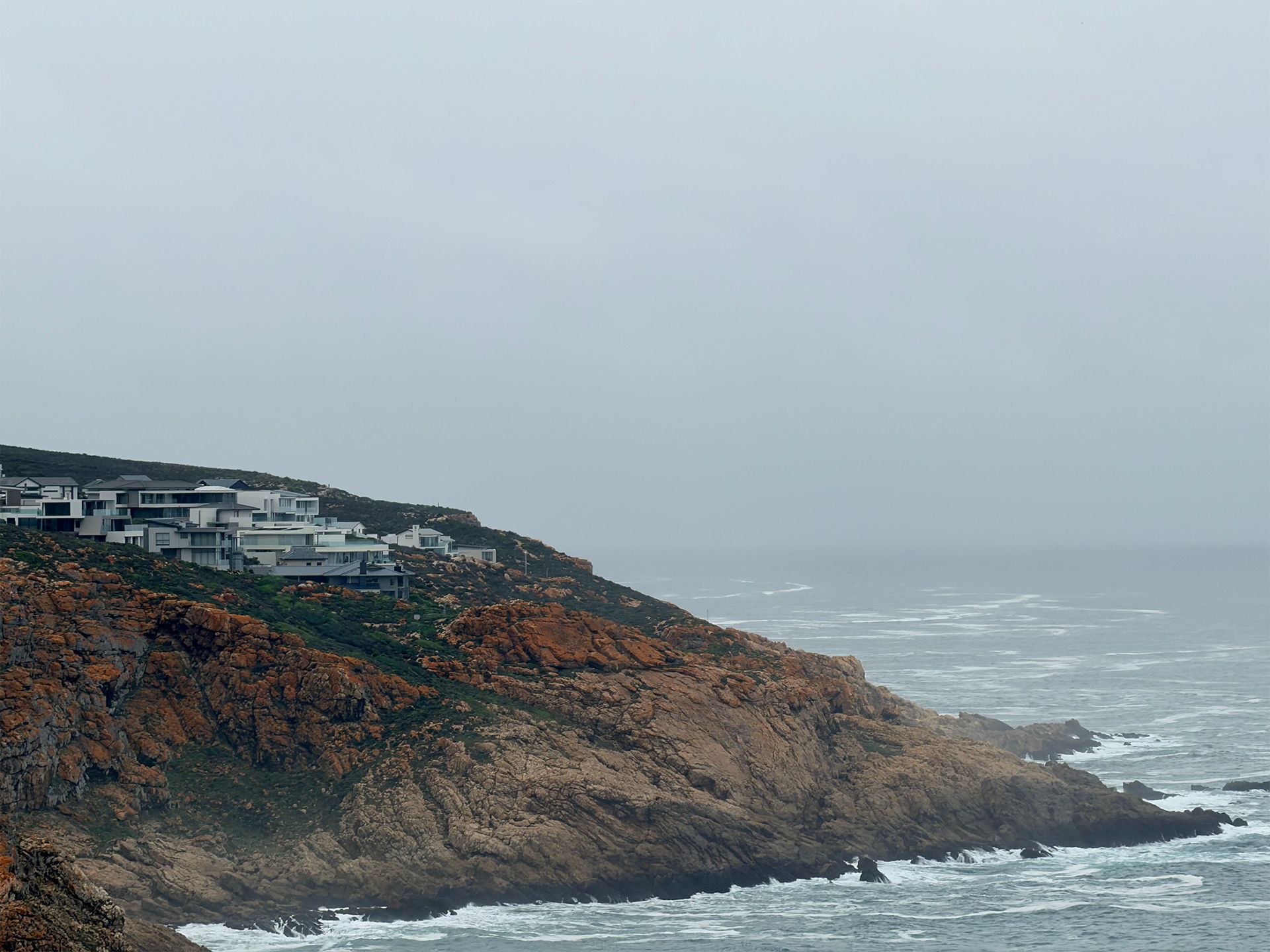 Rocky coastal cliffs with houses under a gray, misty sky beside rough ocean waves.