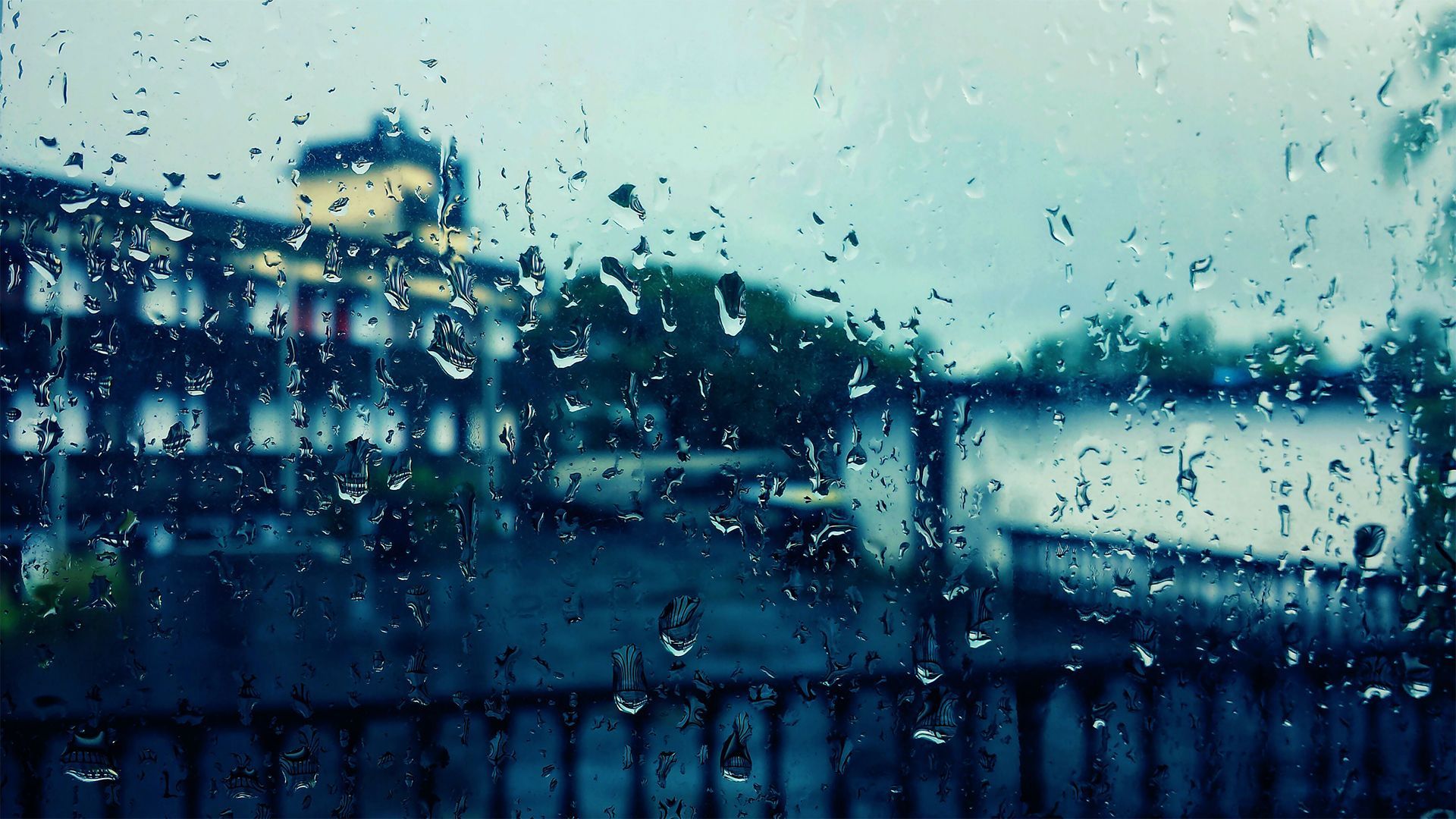 Rain-covered window overlooking blurred blue buildings and a bridge in the distance.