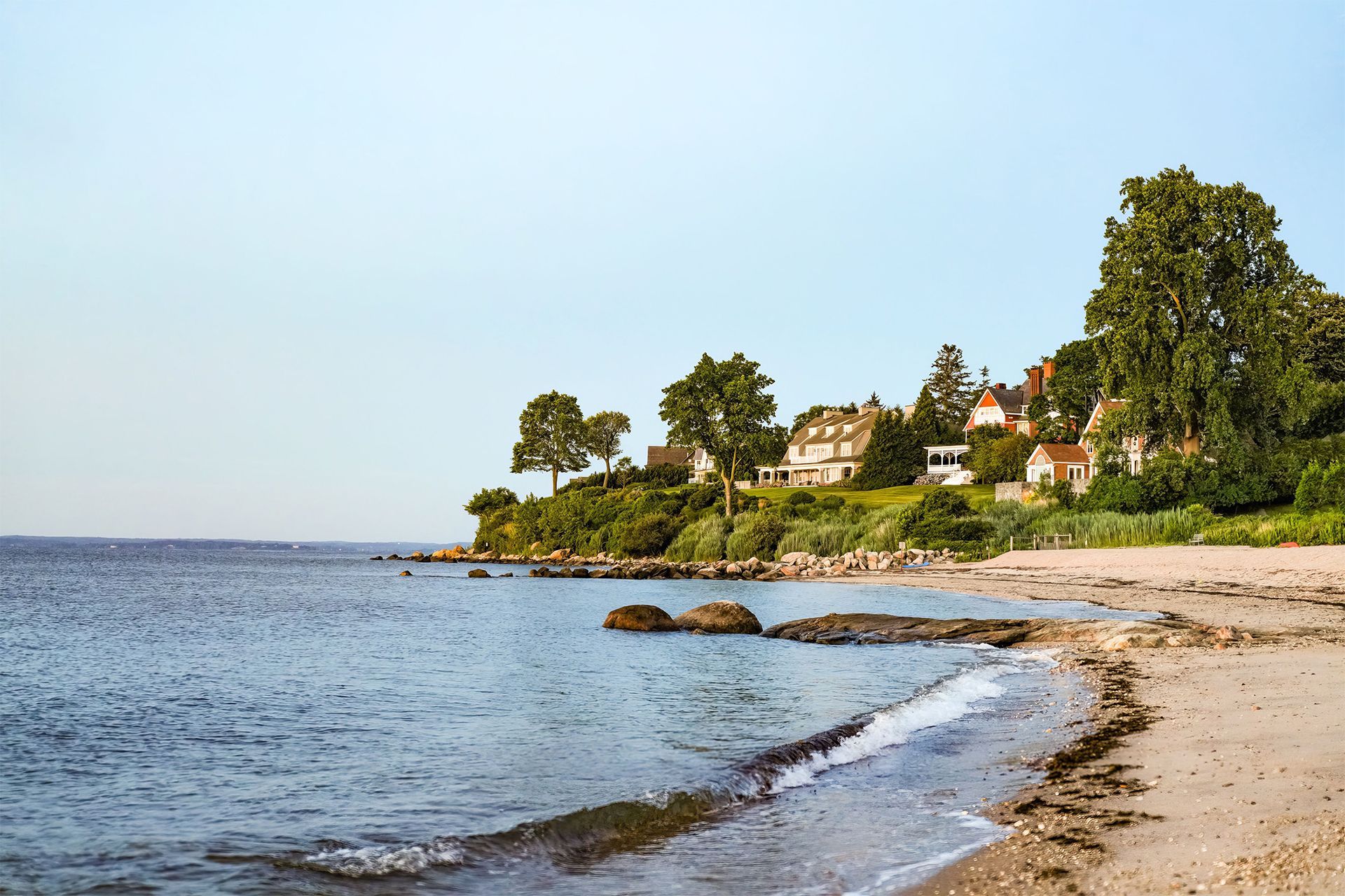 Sandy beach with gentle waves, rocky shoreline, trees, and houses along the coast under a clear sky.