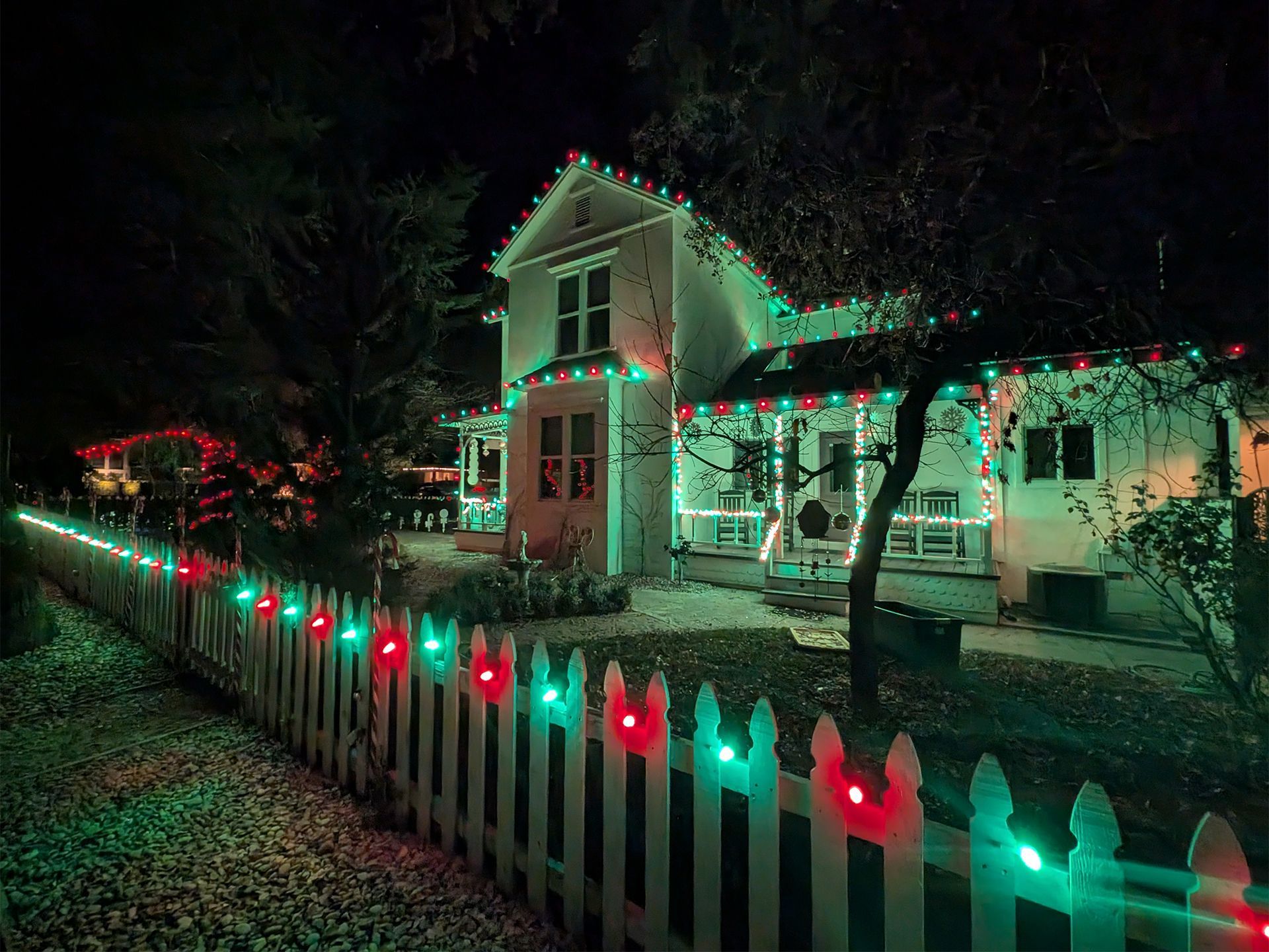 House and white picket fence lit with red and green Christmas lights at night.