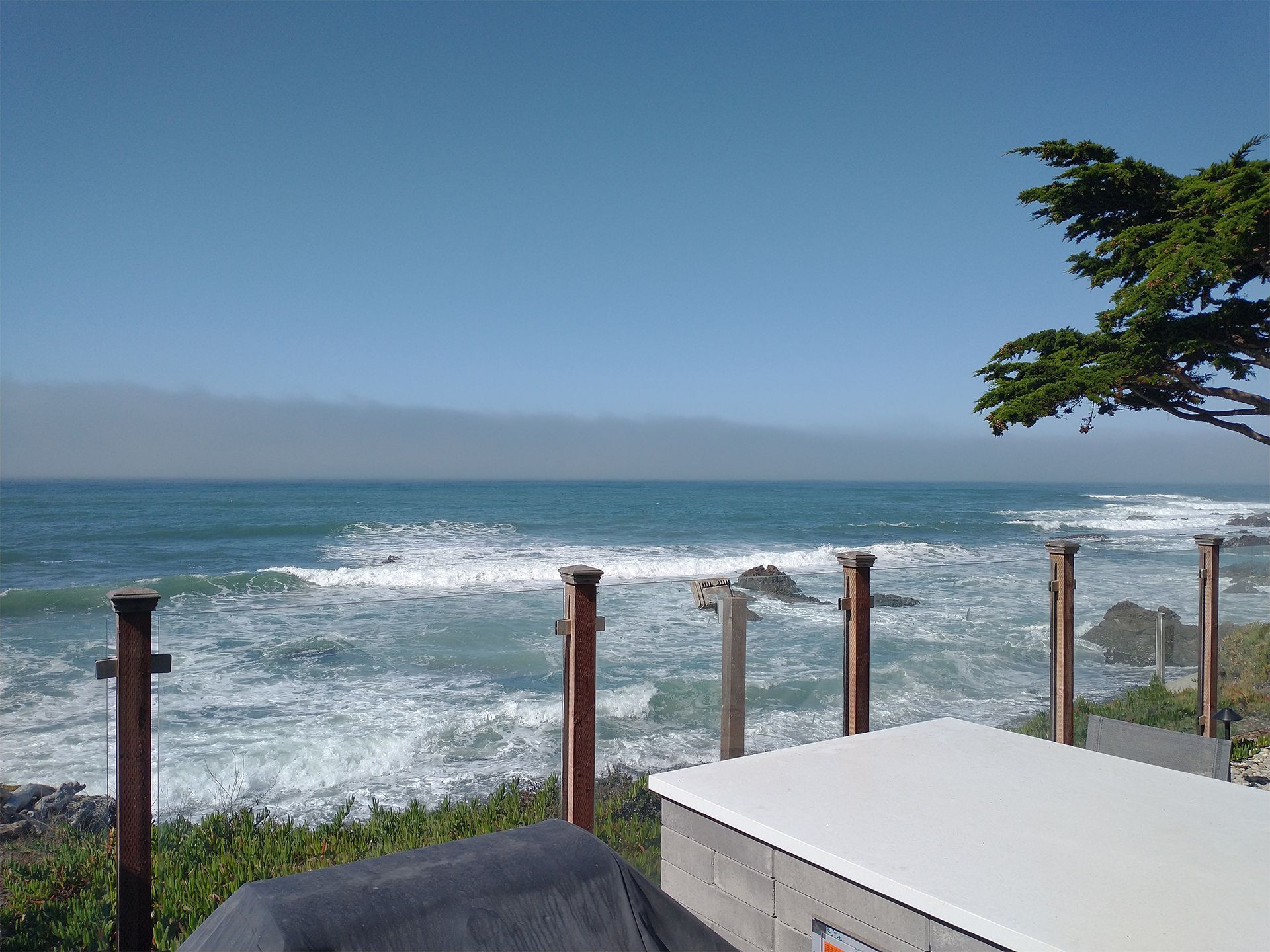 Ocean view from a seaside patio with glass railing, white table, and a tree branch overhead.