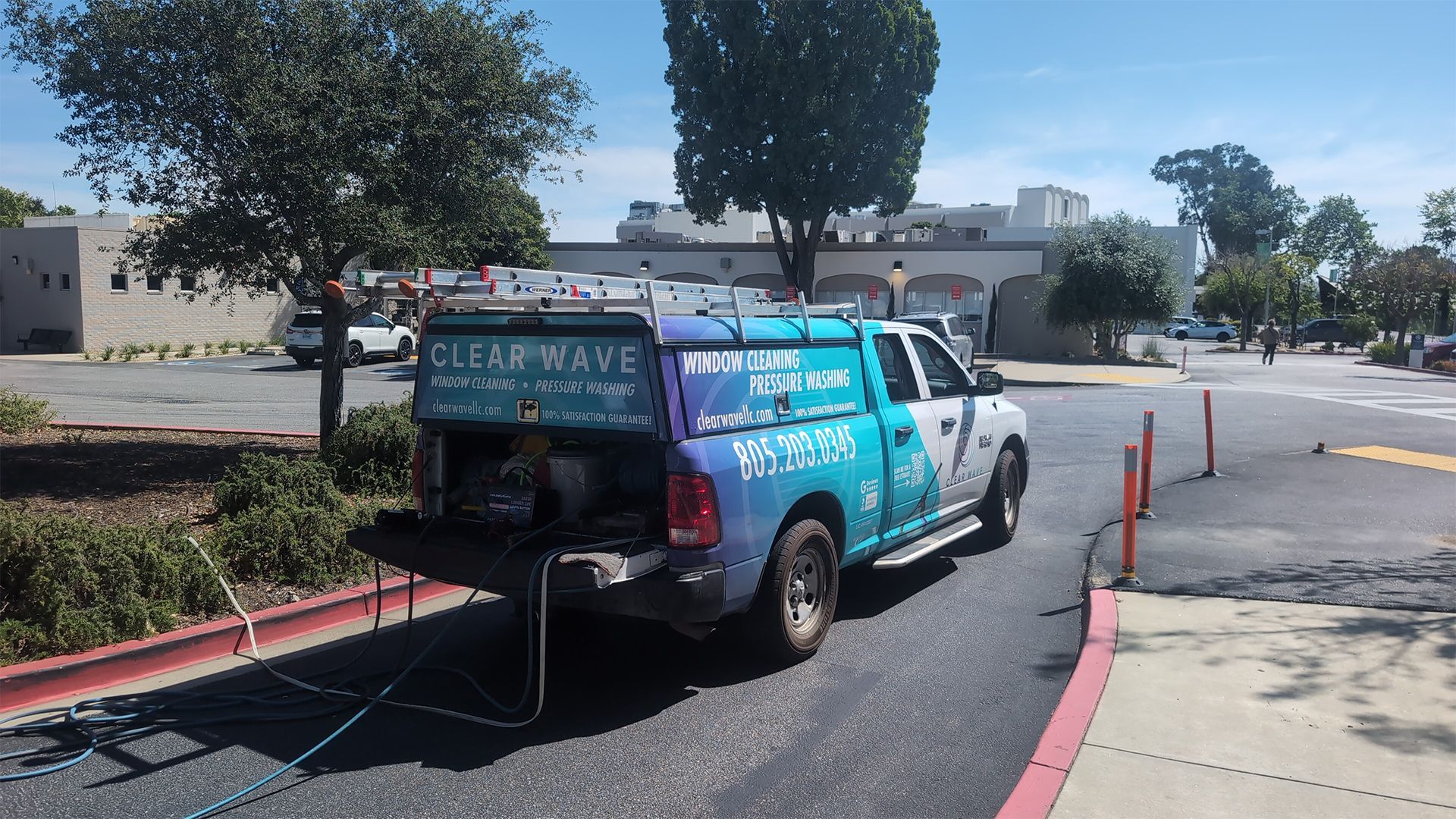 Blue utility truck parked by a curb with orange cones in a parking lot.