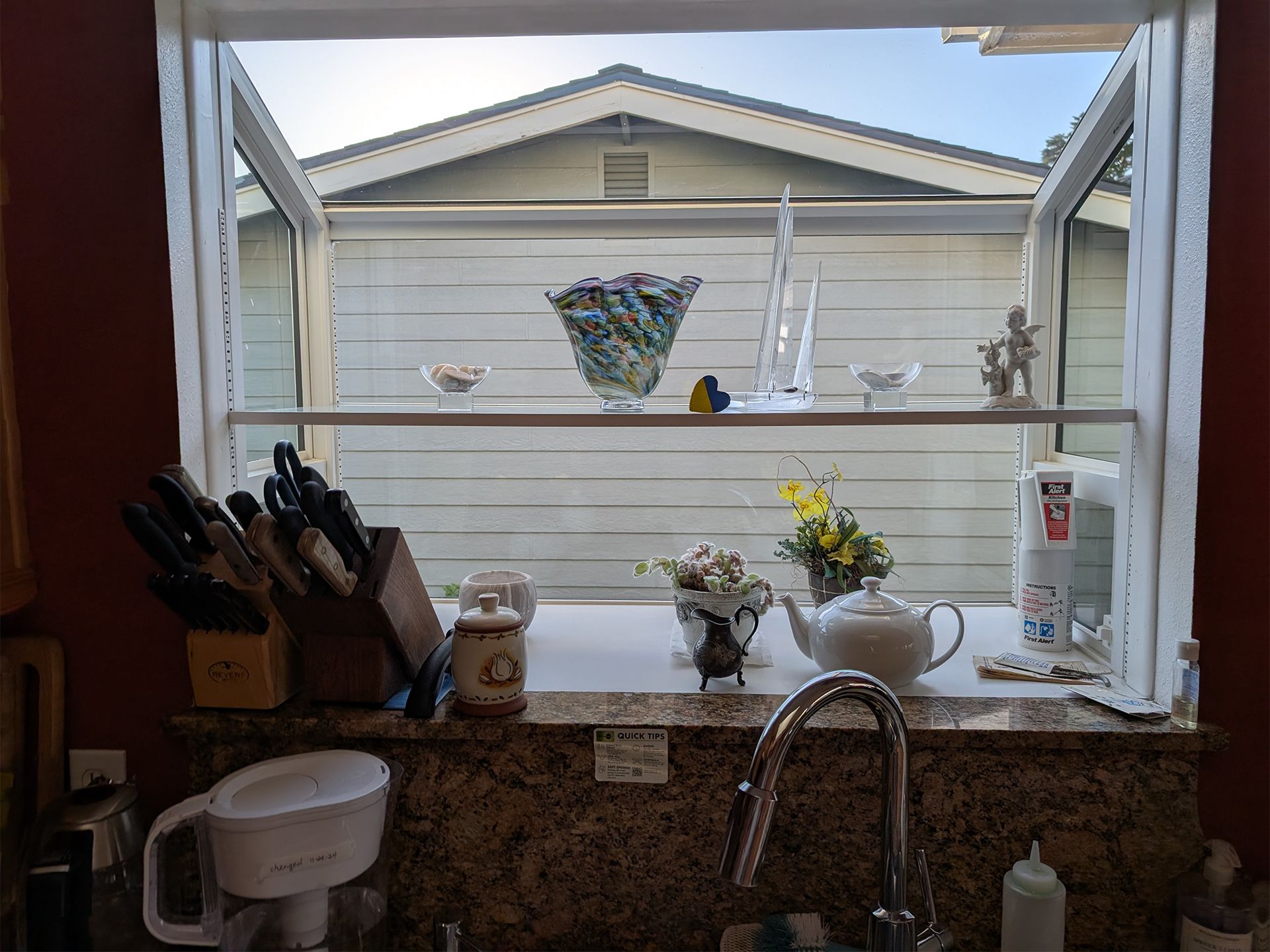 Kitchen sink looking out a window to a backyard fence, with potted flowers on the sill and a knife block on the counter.