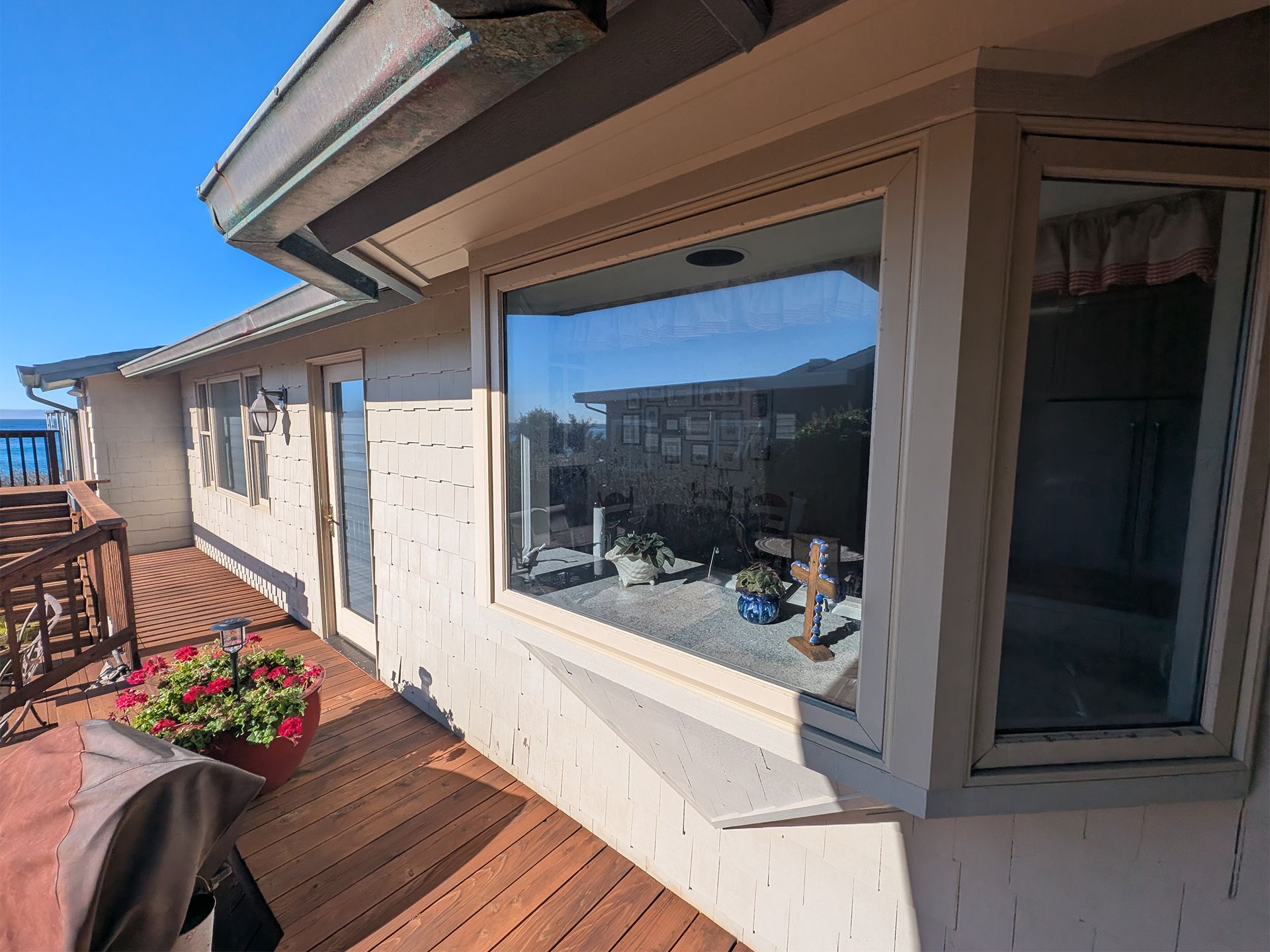 Outdoor wooden deck beside a white house, with a large window and potted flowers in bright sunlight.