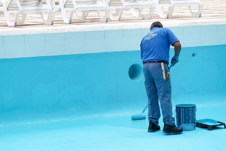 Worker painting a bright blue empty swimming pool with a roller and bucket nearby