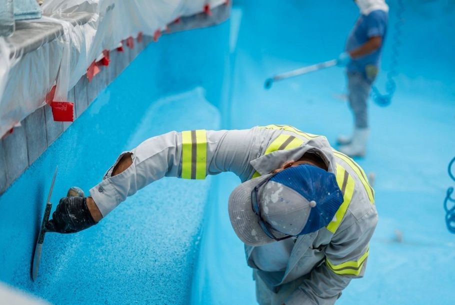 Worker in safety gear painting a bright blue wall in a pool or tank area