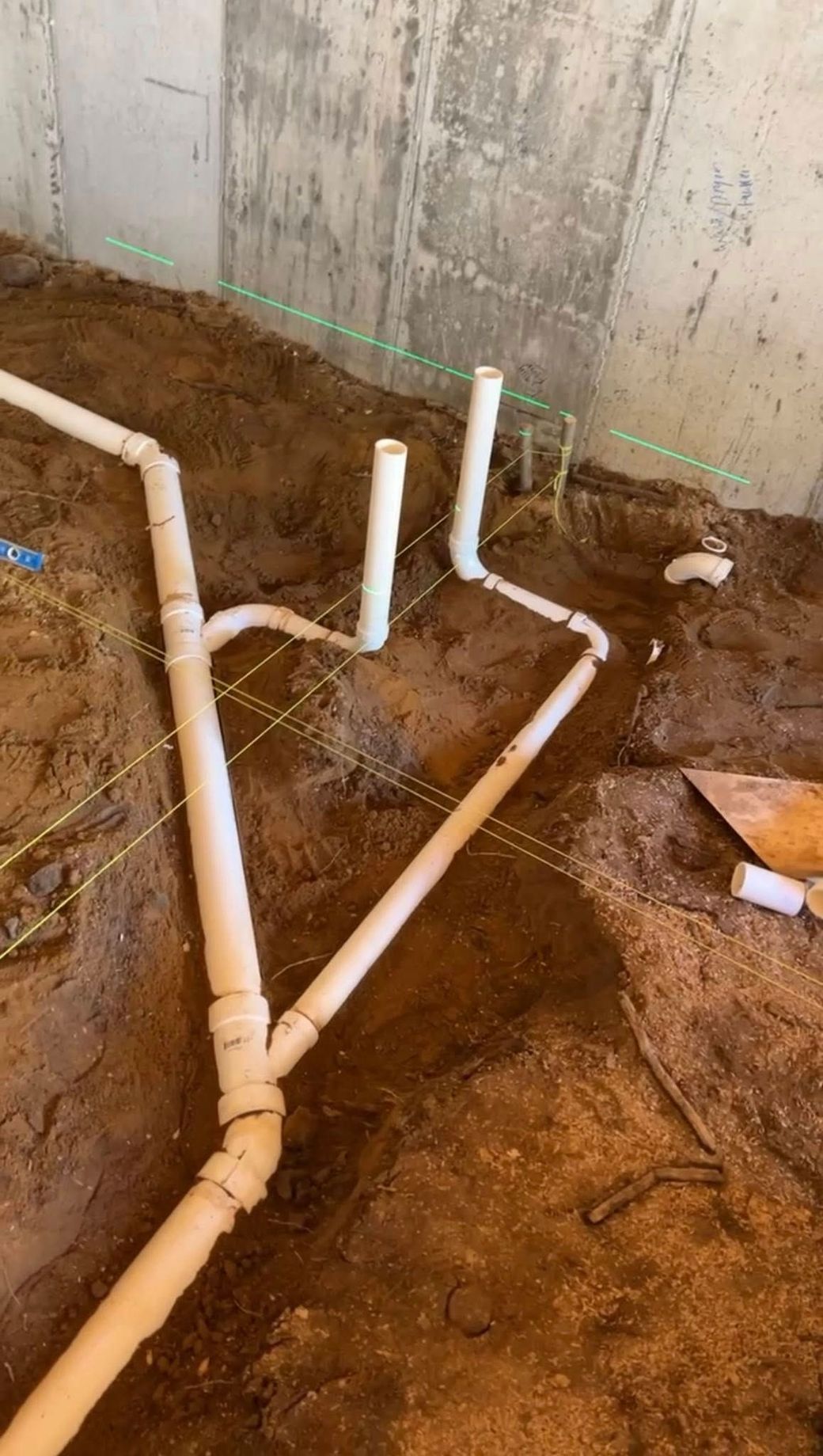 White PVC plumbing pipes installed in a dirt trench, showing a Y-shaped connection and two vertical riser pipes.
