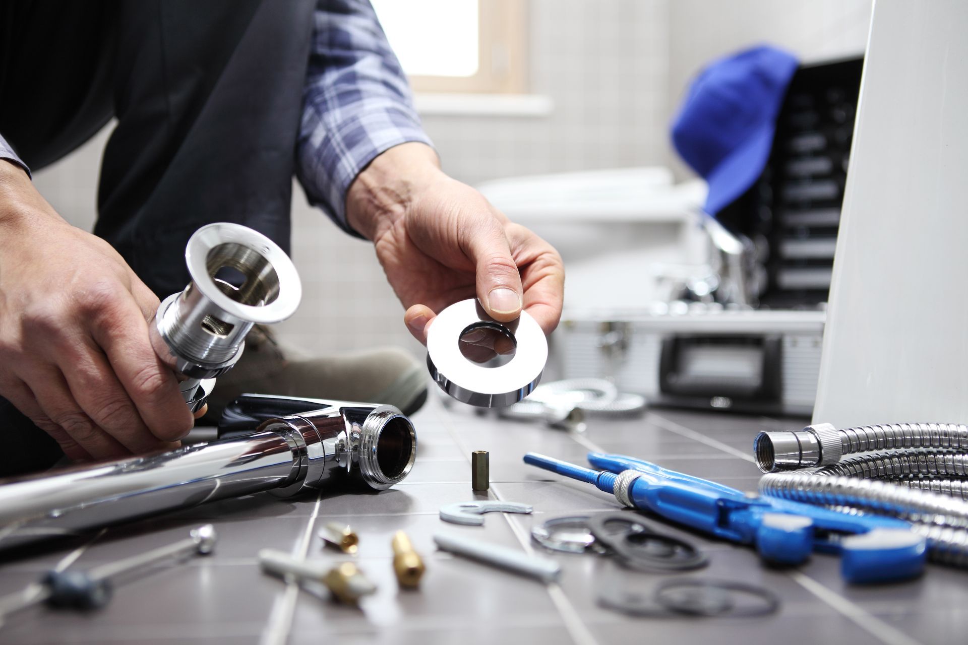 A plumber holding a metal pipe component over a tiled floor with various plumbing tools spread out nearby.