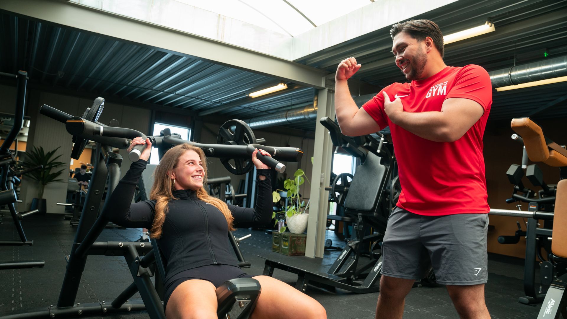 Een vrouw tilt gewichten in de sportschool, ze lacht en er is een instructeur die haar biceps traint.