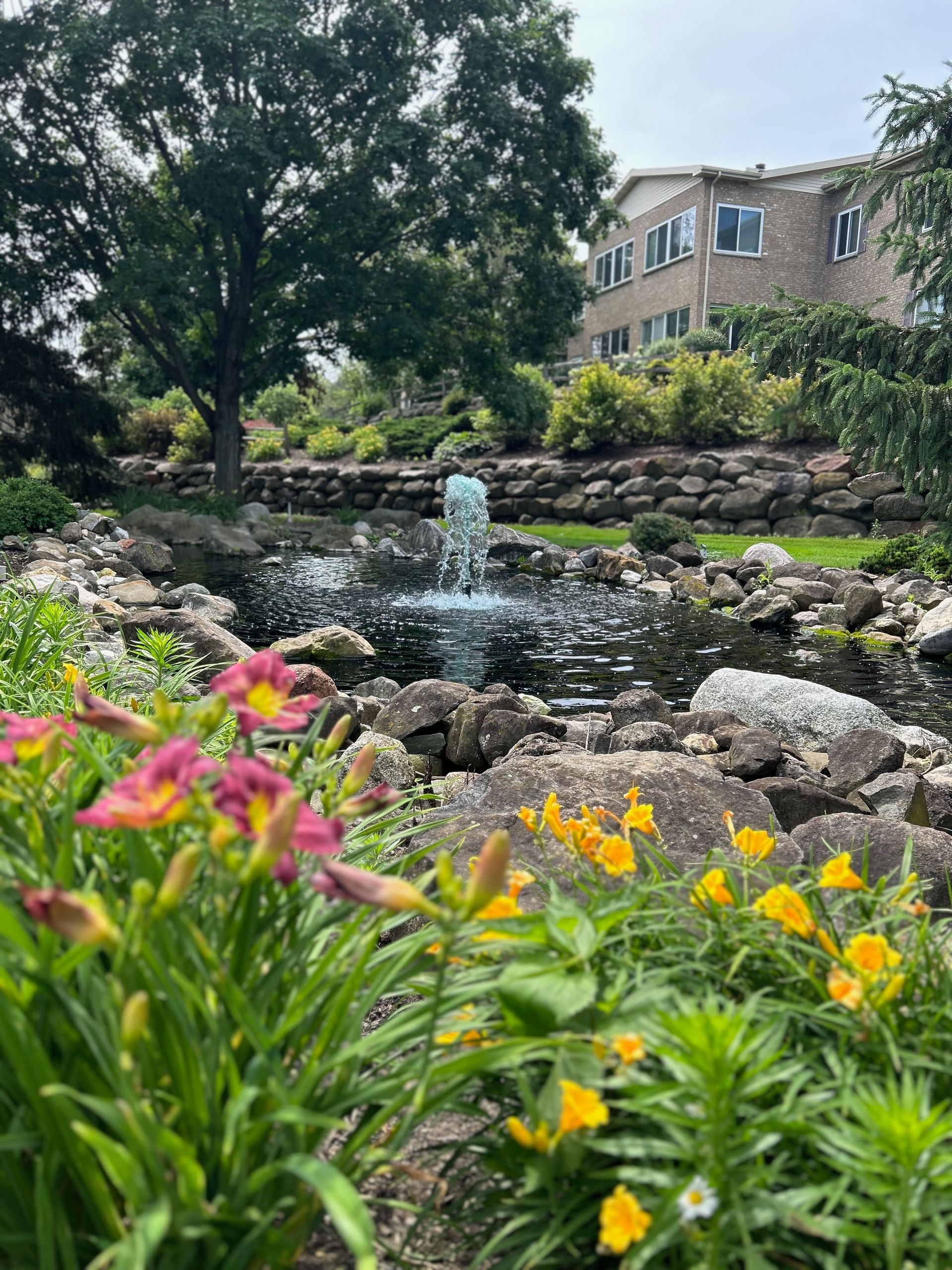 A fountain in a garden with flowers in the foreground and a building in the background.