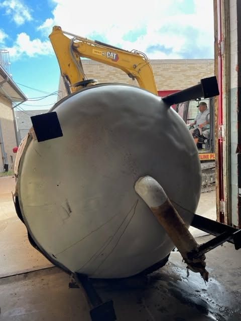 Large gray cylindrical tank being hoisted by an excavator at a construction site.