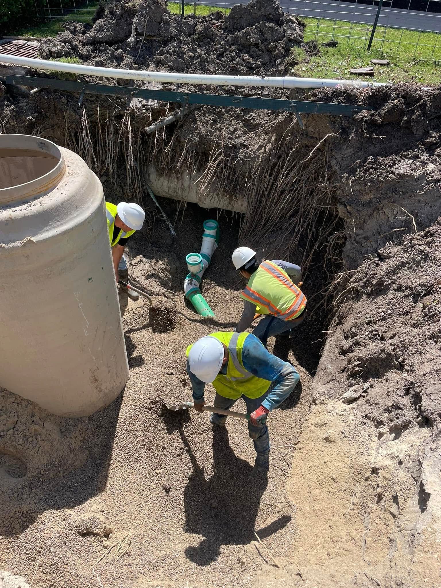 Workers in neon vests digging a trench beside a large concrete pipe in bright sunlight