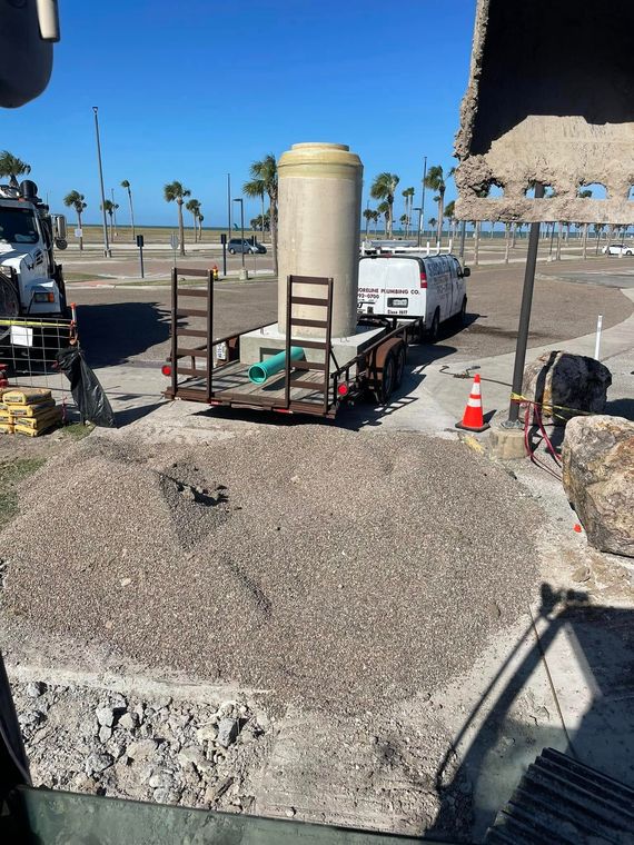 Beachside construction equipment and supplies beside a stone wall, with ocean and palm trees in the background.