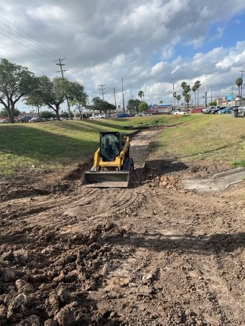 Small excavator working in a muddy drainage ditch beside a road under a cloudy sky