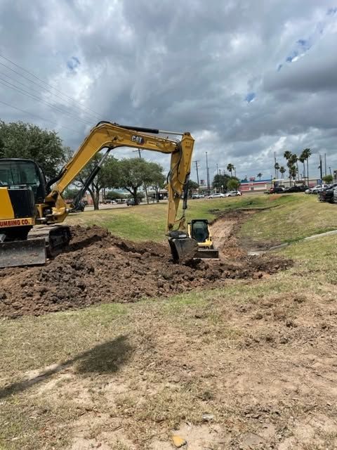 Yellow excavator digging a trench beside a grassy roadside under cloudy skies
