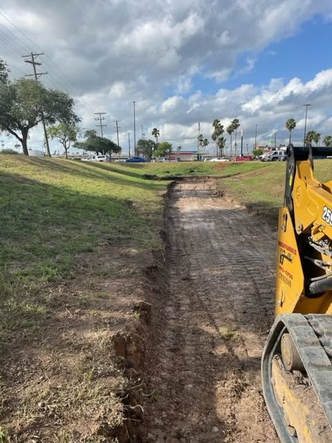 Dirt path being dug beside a grassy slope, with part of a yellow excavator visible on the right.