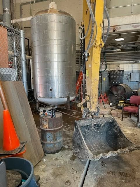 Industrial workshop with a large metal tank, yellow hoist, and wheelbarrow on a wet concrete floor.