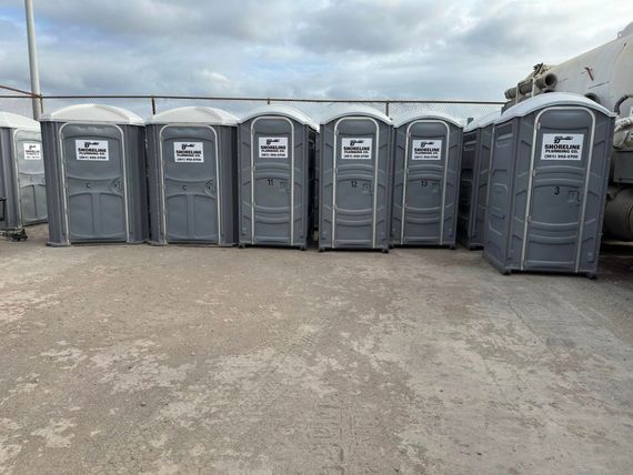 Row of gray portable toilets lined up outdoors on a gravel lot under a cloudy sky