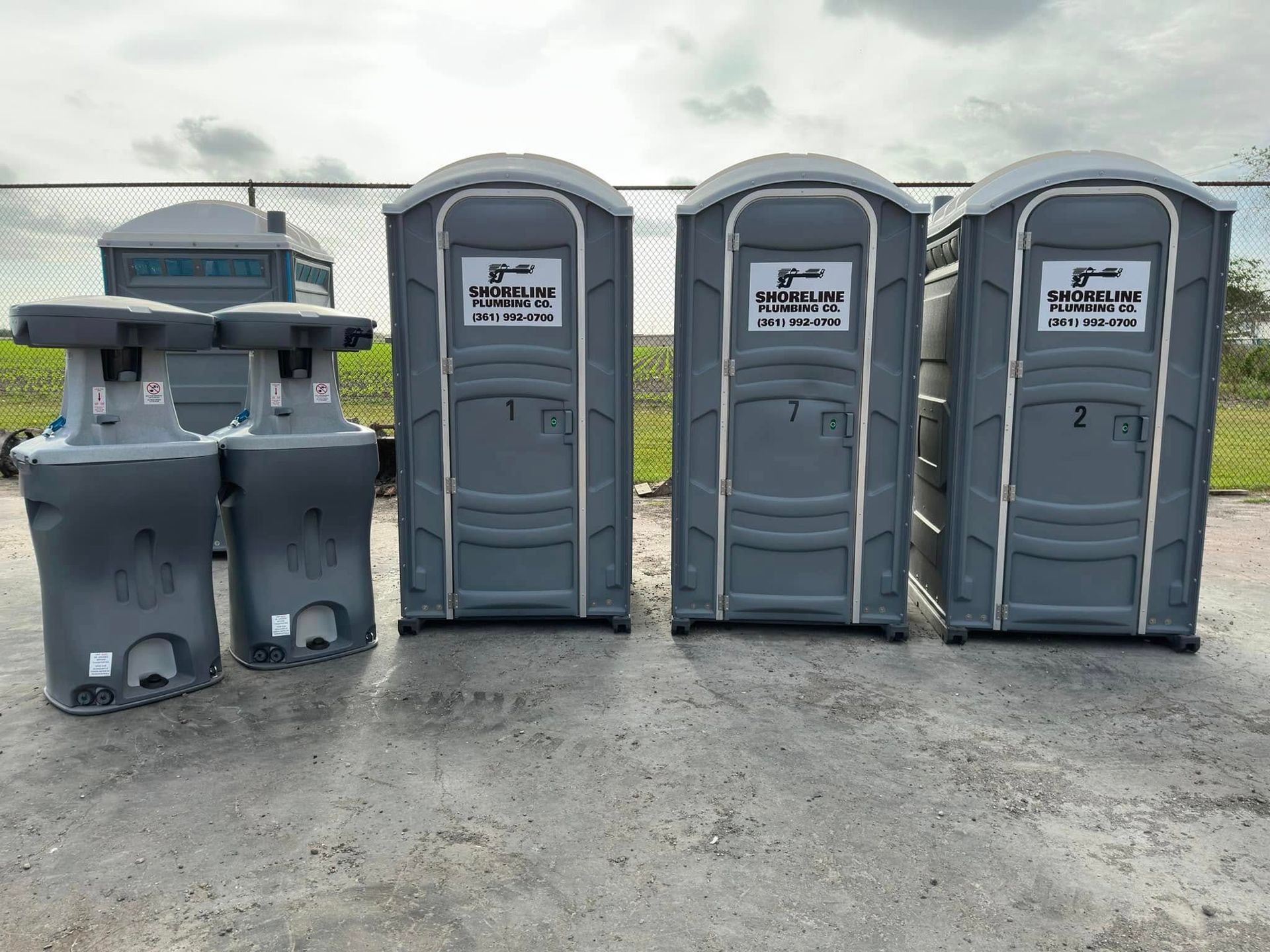 Four portable toilets and two gray handwashing stations on a gravel lot outdoors