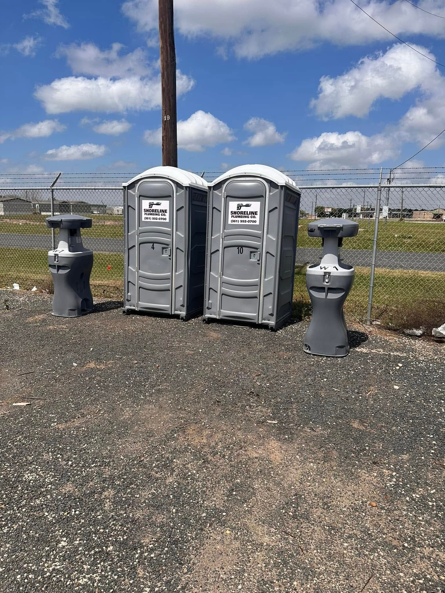 Two portable toilets on a gravel lot beside a chain-link fence under a blue sky