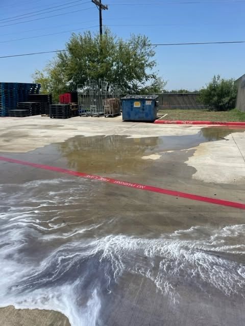 Wet concrete lot with puddle runoff, blue dumpster, and trees under a clear sky
