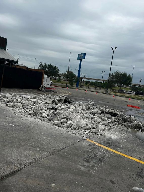 Rubble and broken concrete scattered across a parking lot under a cloudy sky, with a distant sign and light poles.