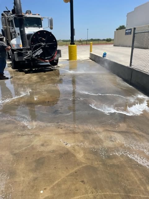 Black truck spraying water on a wet concrete lot beside a chain-link fence and yellow bollard.