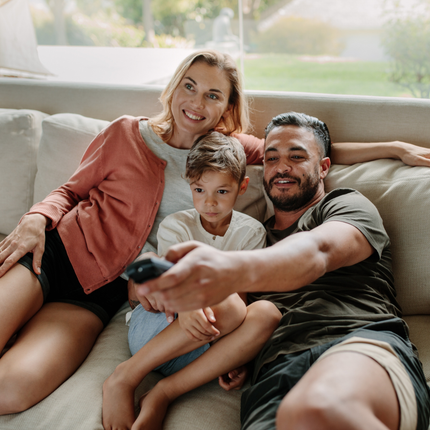 Family of three on a couch, watching TV; the man holds a remote control. Woman smiles, child looks serious.