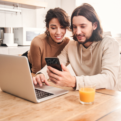 A couple in a kitchen looks at a phone and laptop. The woman smiles as the man holds the phone. 