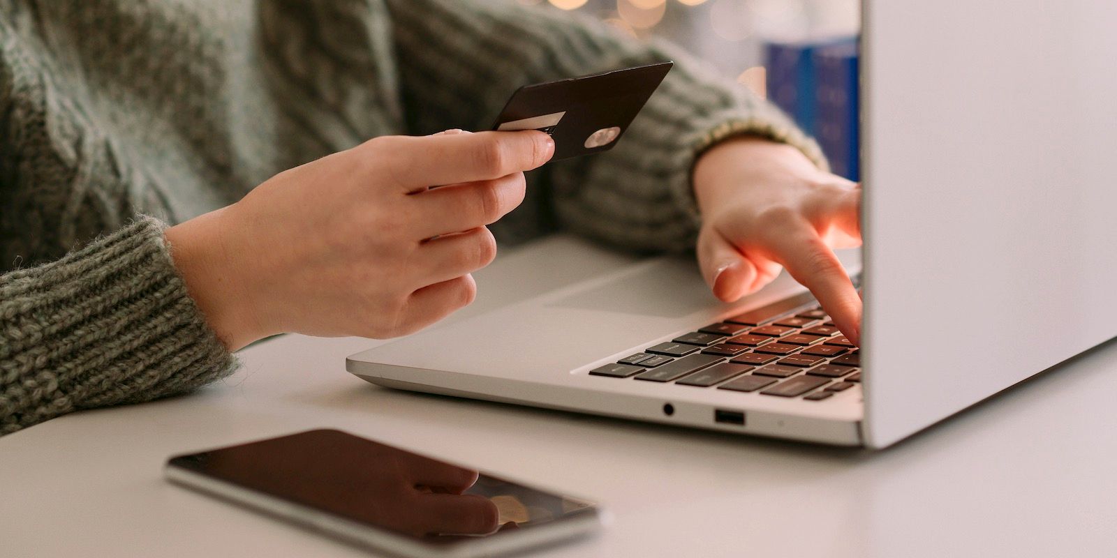 Person using a laptop with a credit card in hand and a phone on the table, presumably for online shopping.