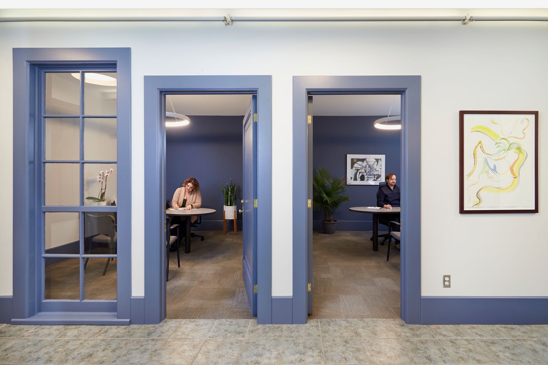Three office doorways with blue trim and gray walls, with two people working inside. One door is glass.