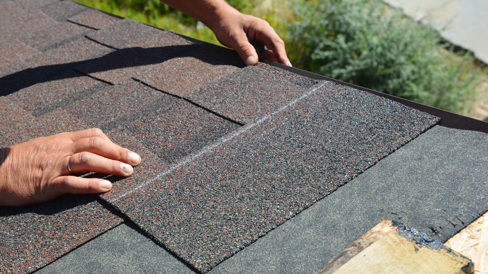 Hands installing a dark-colored asphalt shingle on a roof.
