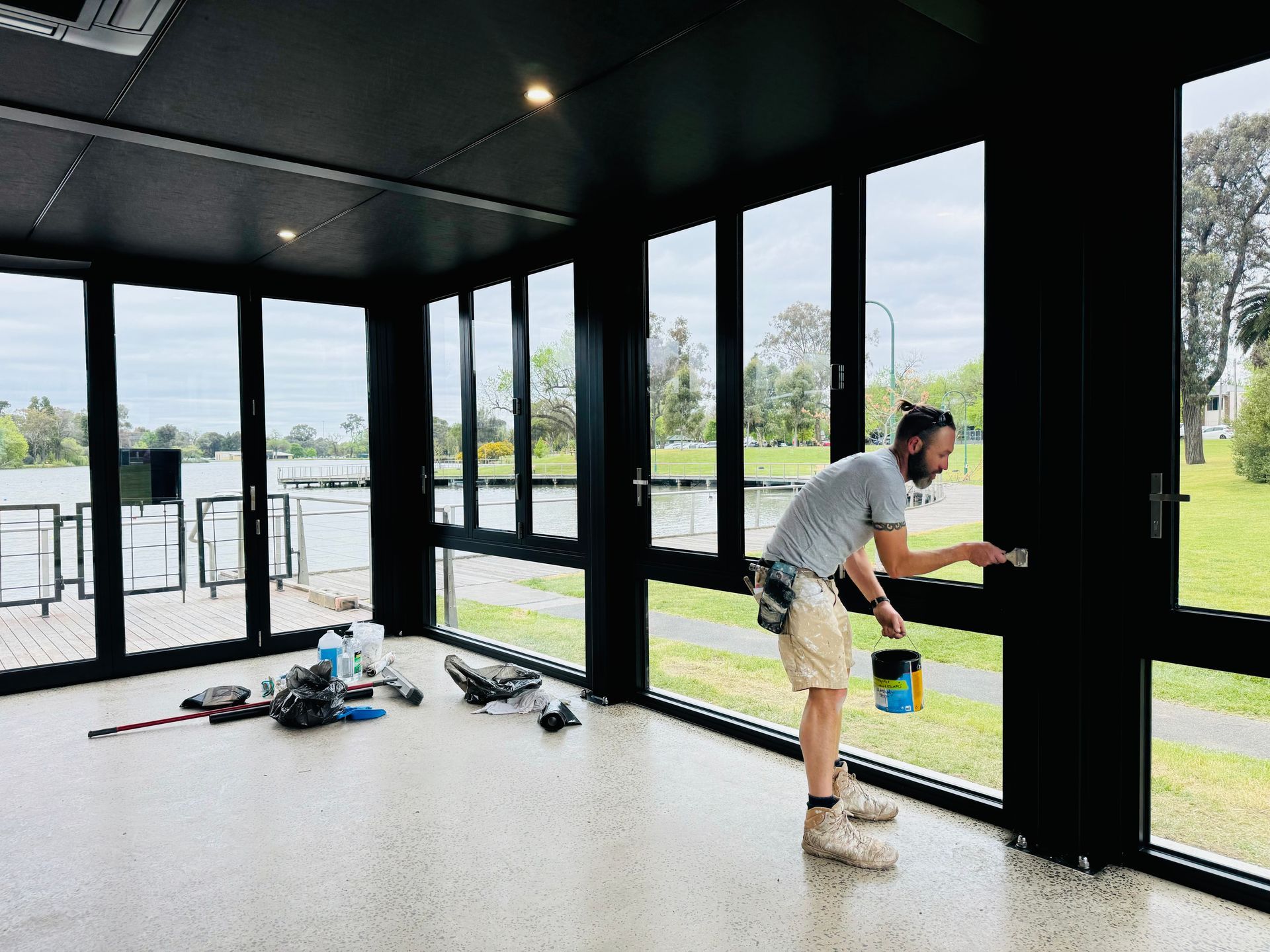 Man painting black window frame indoors with waterfront view — Crampton Painting & Decorating Pty Ltd In Maiden Gully, VIC