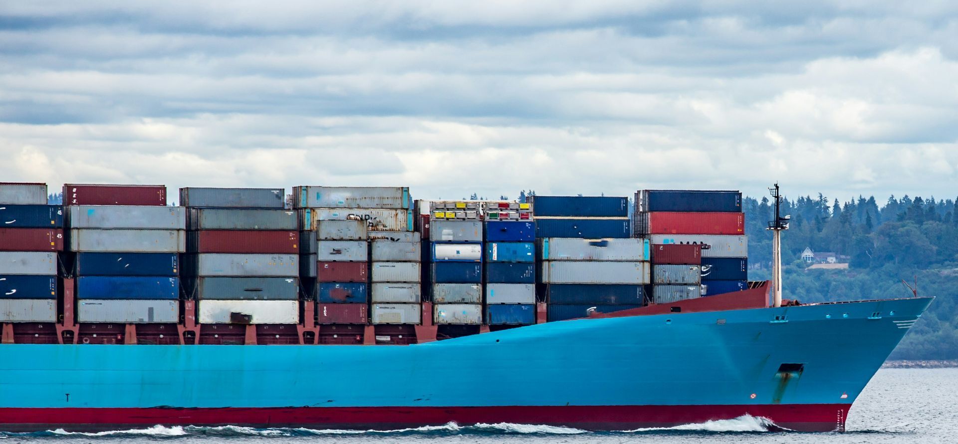 A large teal cargo ship carrying stacked shipping containers on a body of water.