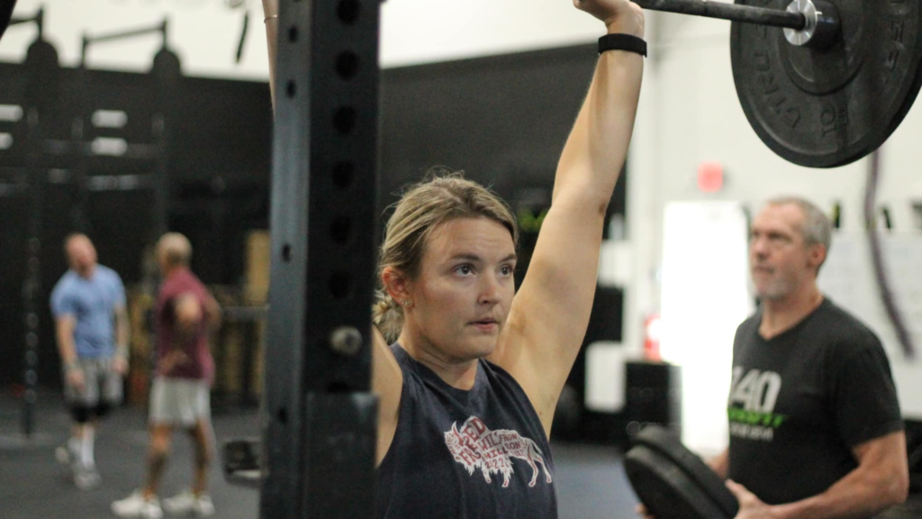 A focused individual lifts a barbell overhead in a gym setting while a trainer watches nearby.
