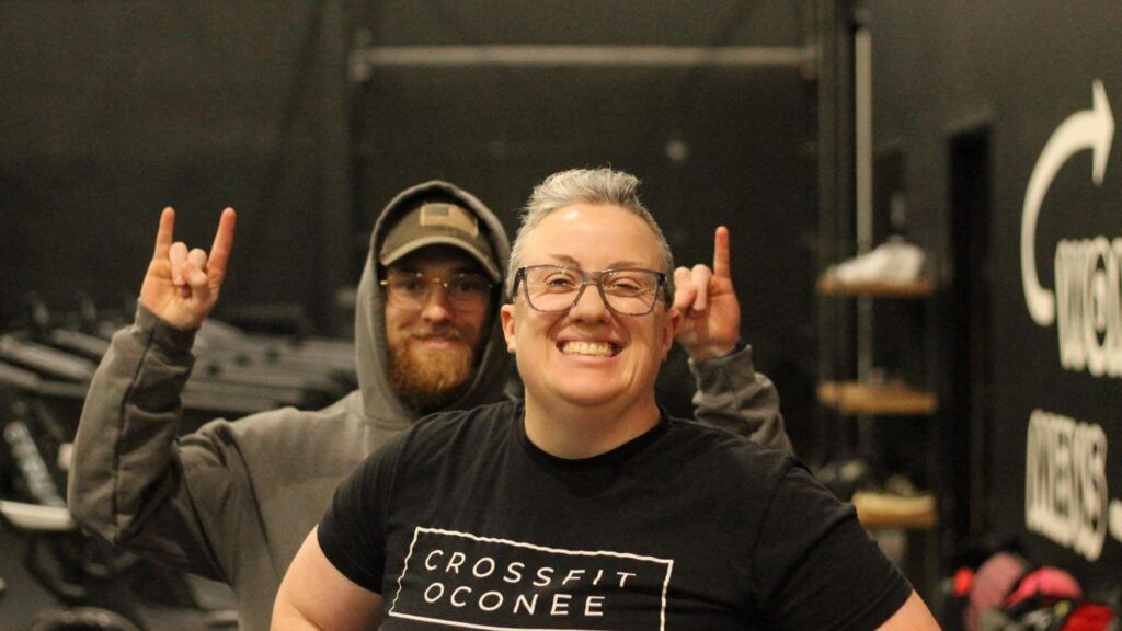 Two people smiling and making hand signals in a CrossFit gym with black walls and equipment in the background.