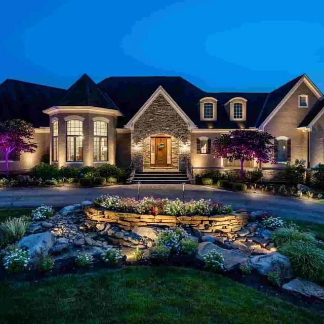 Luxury brick house at dusk with glowing windows, landscaped garden, and illuminated stone walkway