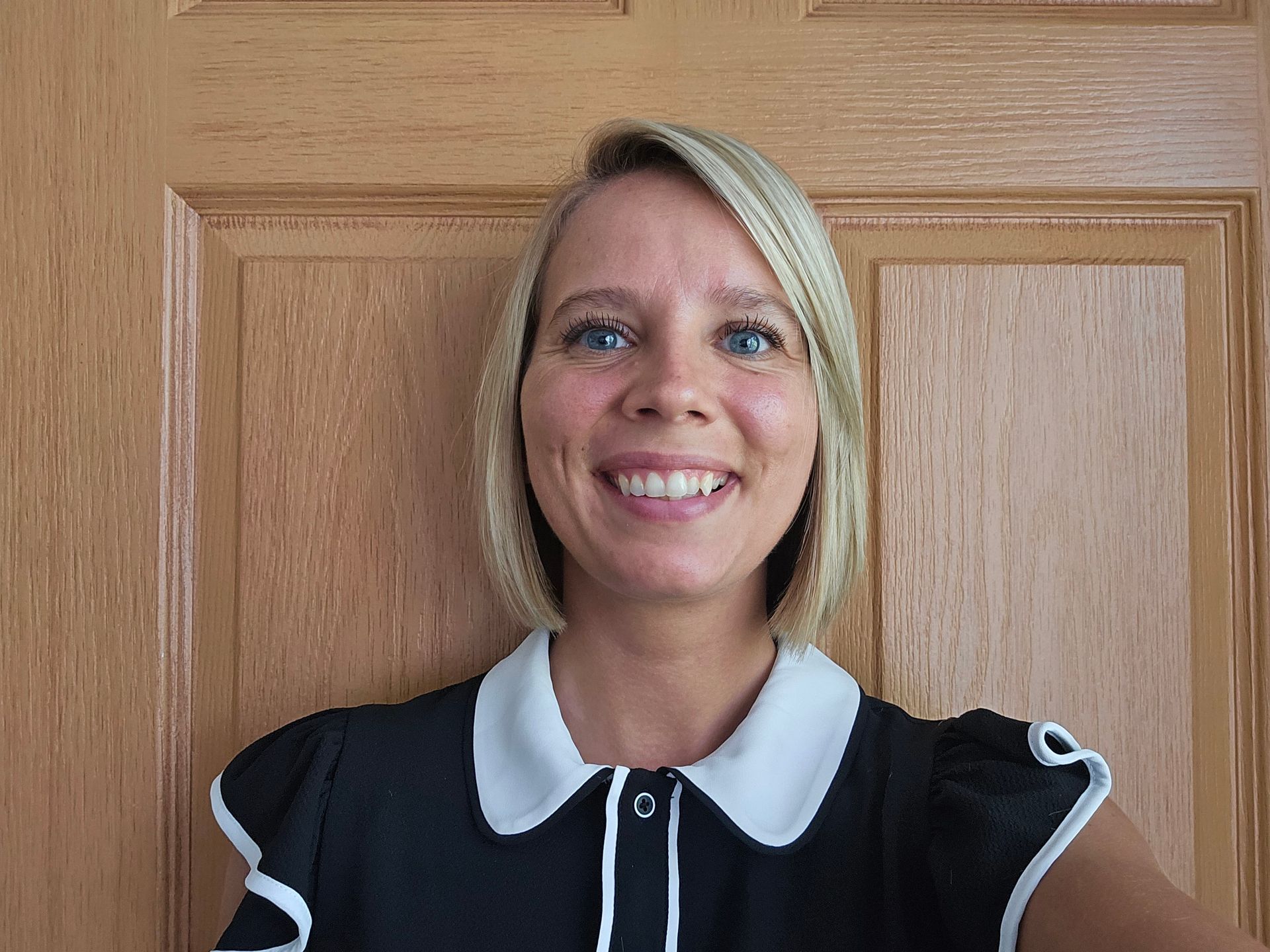 Smiling woman in black uniform with white collar, standing against a wooden door backdrop