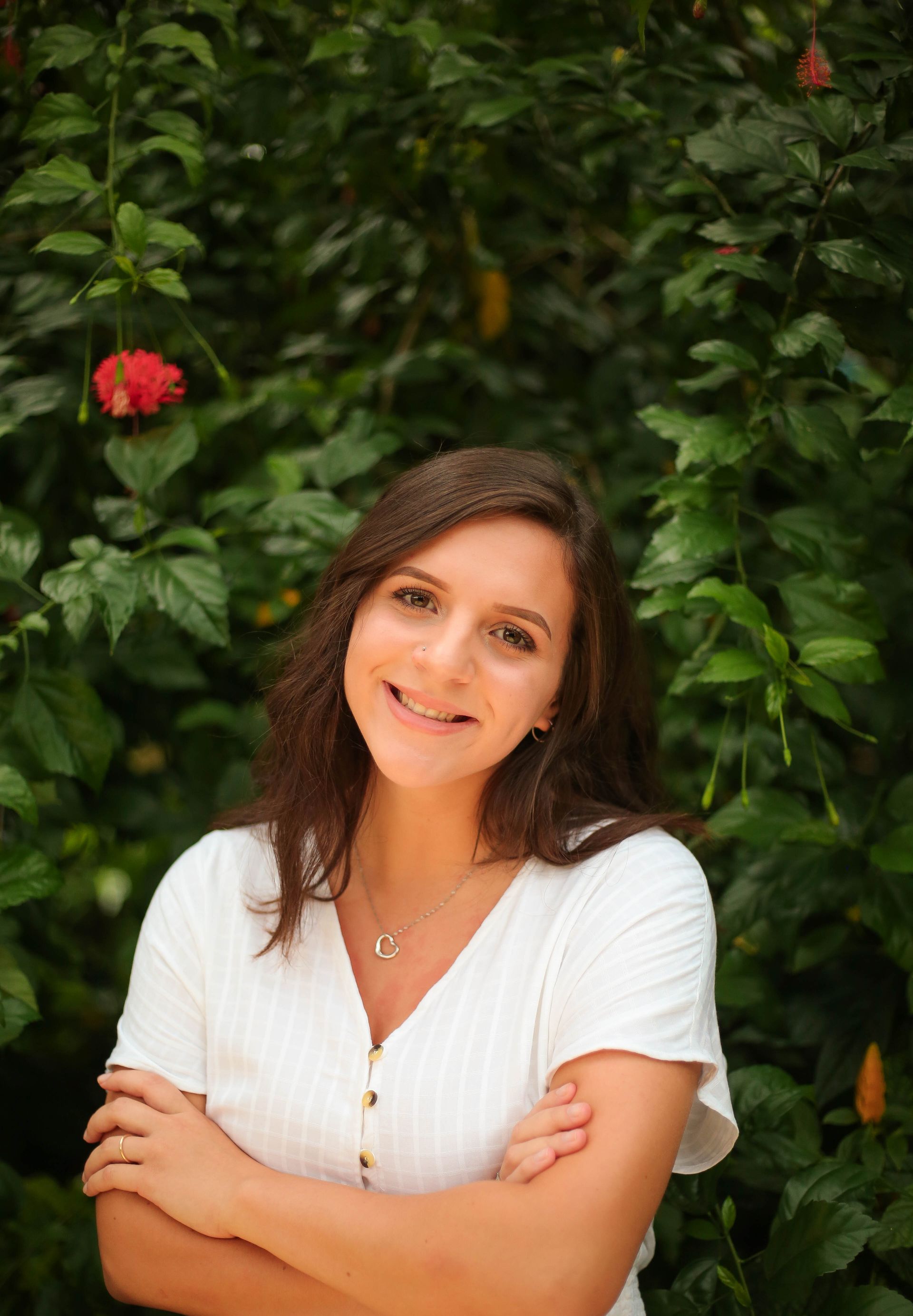 Smiling woman in a white top with arms crossed, standing before lush green foliage and red flowers