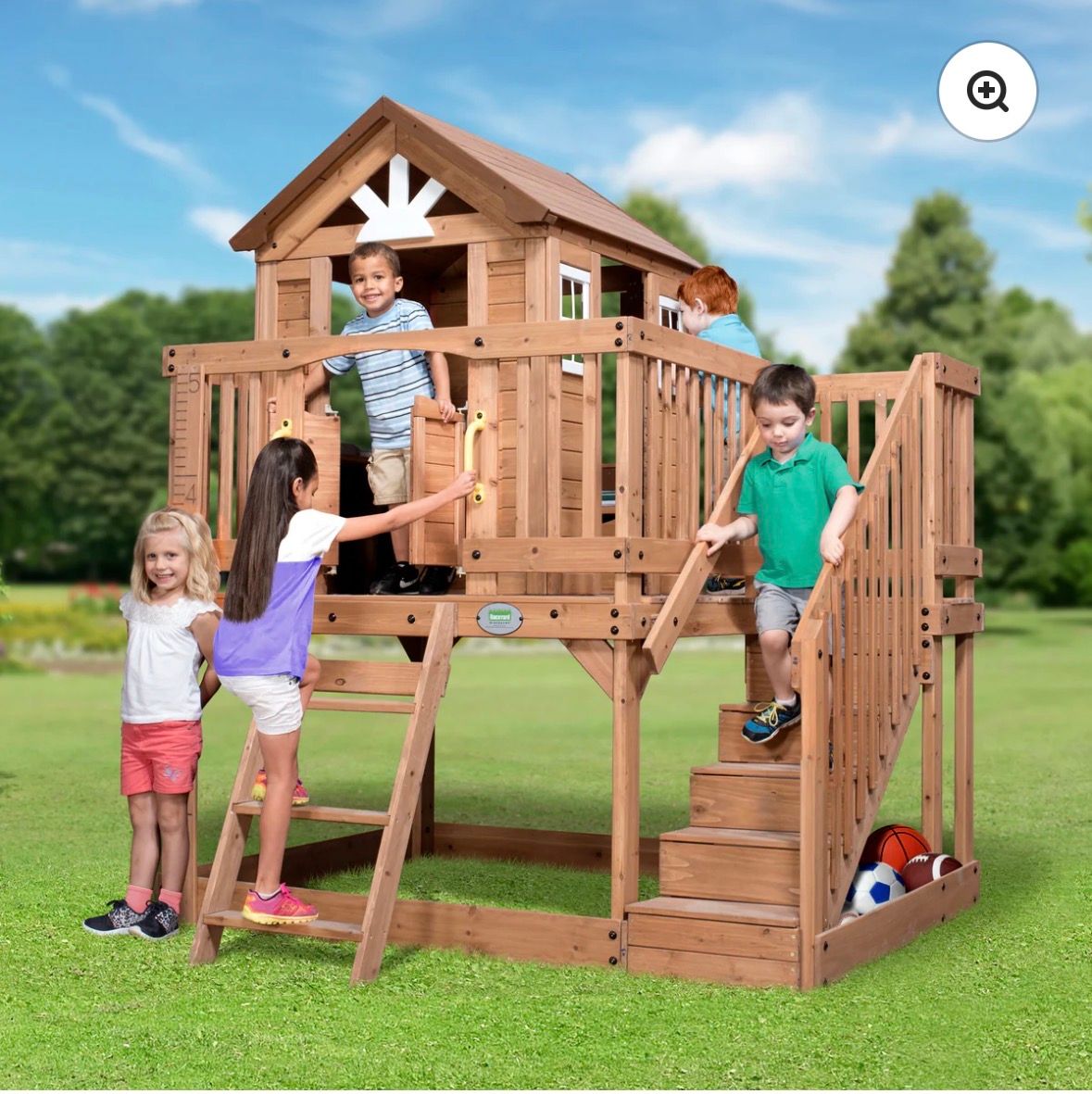 Children playing on a wooden backyard playset with stairs and a slide in a grassy yard.