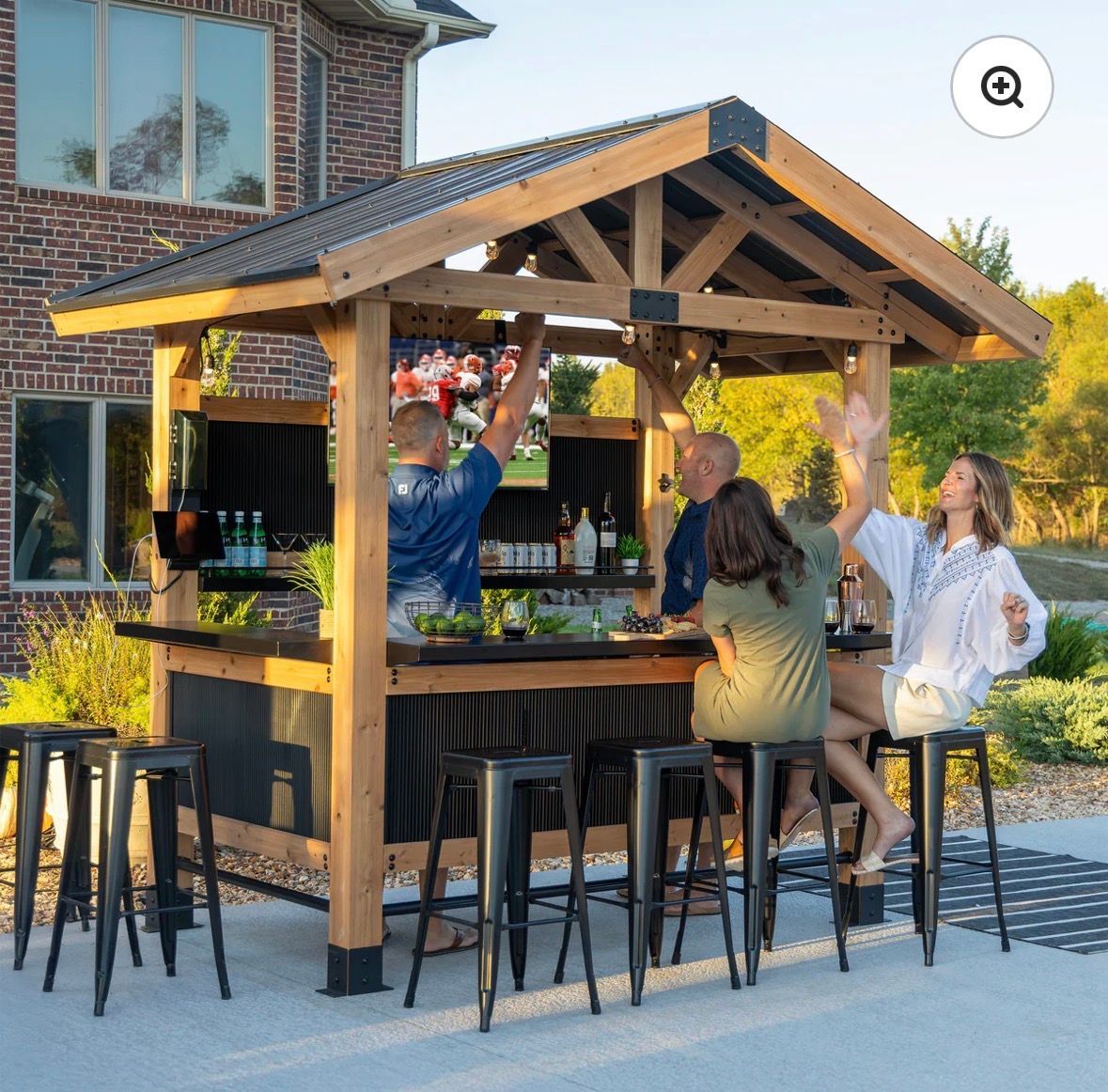 People seated at a wooden outdoor bar under a pavilion, socializing on a patio with stools and drinks.