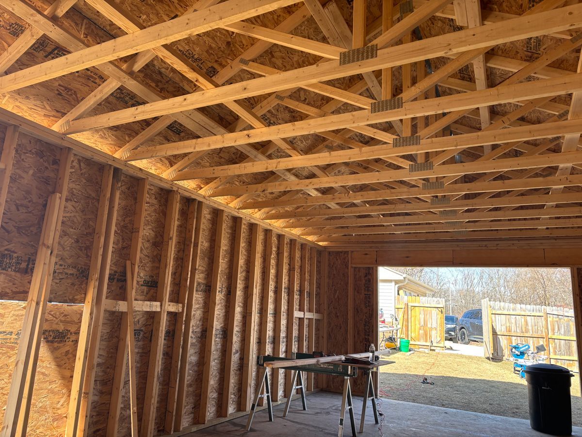 Interior of a wooden-framed construction site with exposed studs and roof trusses opening to a sunlit yard.