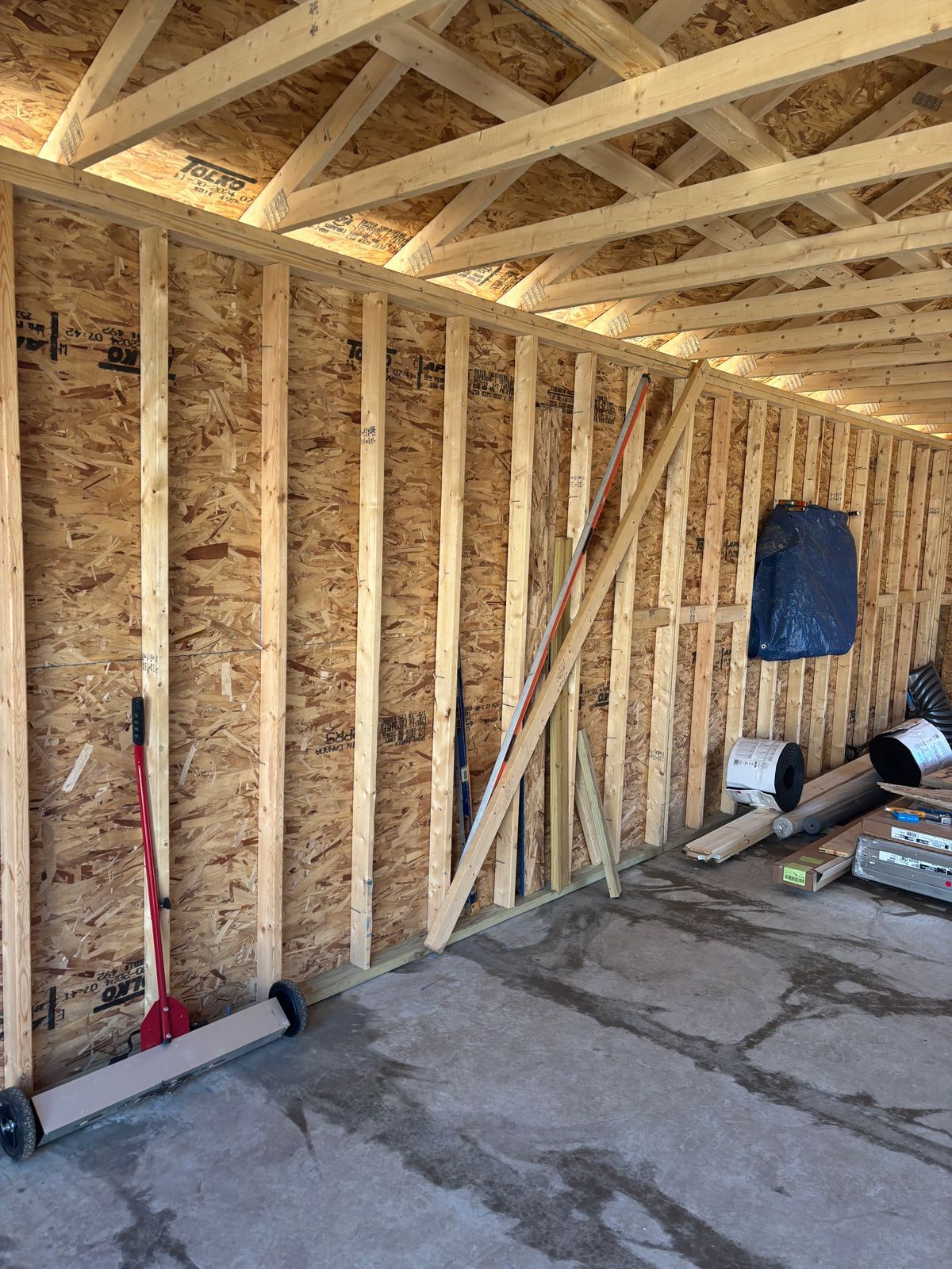 Unfinished garage interior with exposed wood framing, concrete floor, and tools leaning against the wall