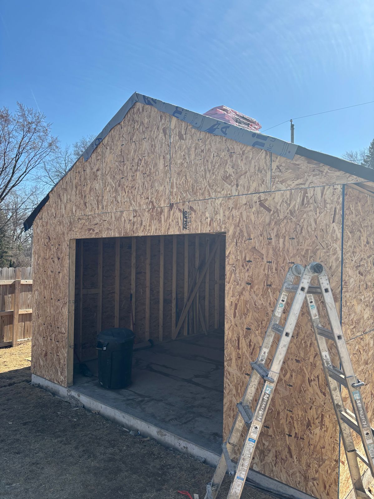 Partially built plywood garage with open doorway and a ladder leaning outside in sunlight