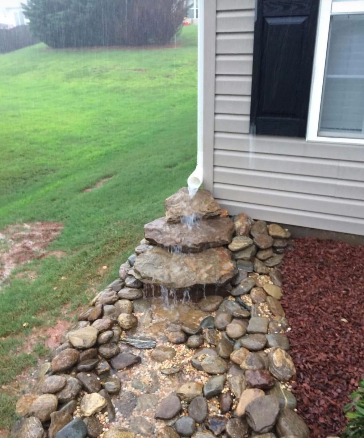 Stacked rock fountain beside a house with water spilling over stone tiers into a small garden bed