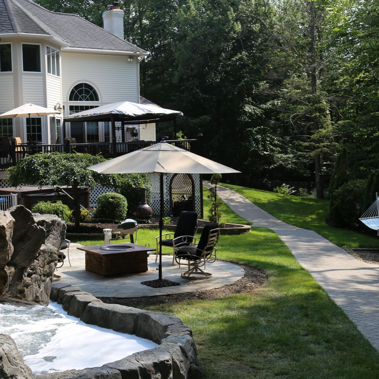 Patio with umbrellas and chairs beside a stone-lined stream and a large house in a wooded yard