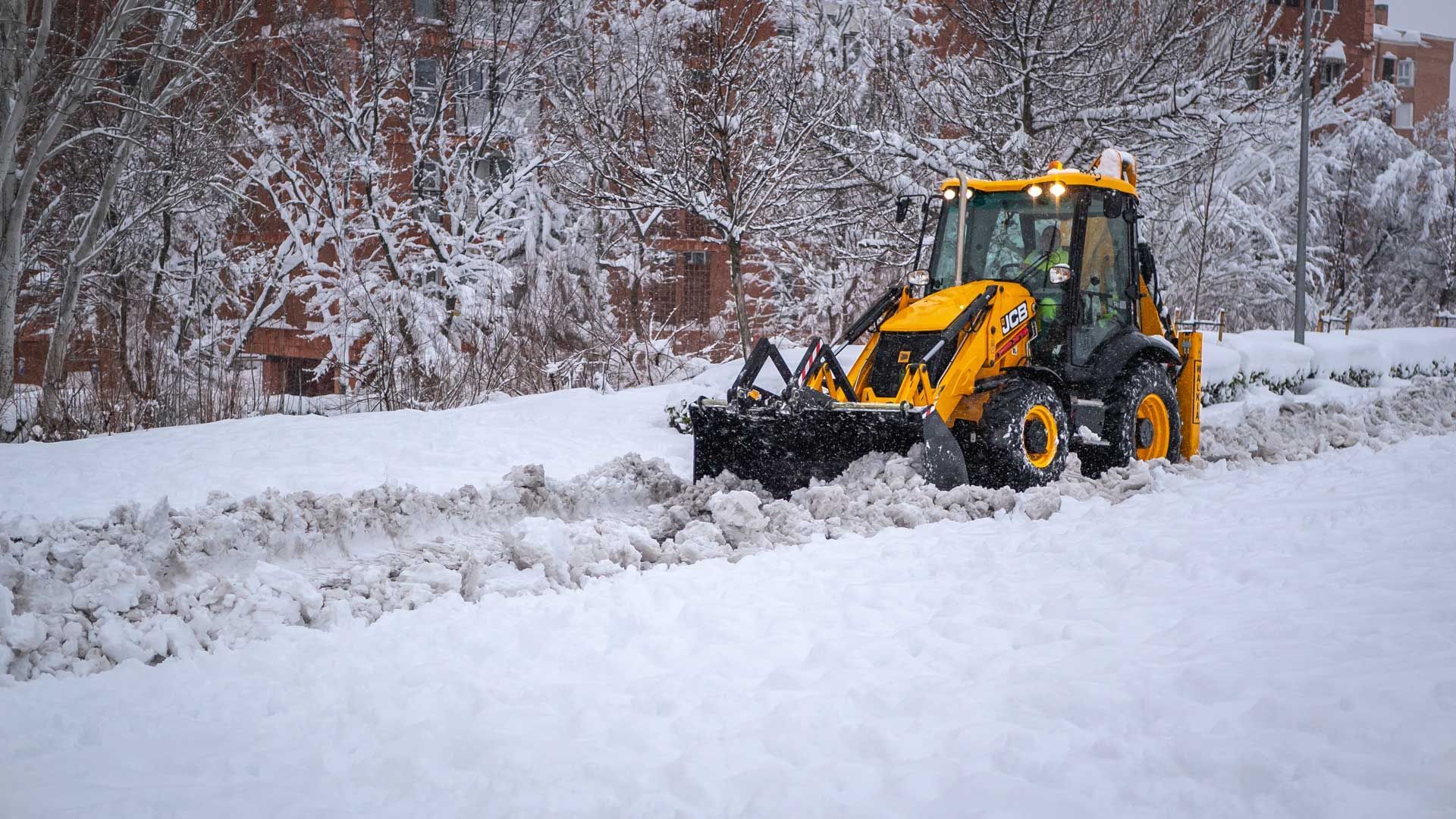 Yellow backhoe clearing snow on a snowy road beside snow-covered trees